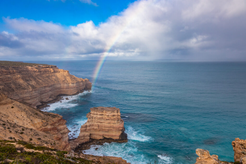 Rainbow stretching across the sky above Island Rock and the rugged Kalbarri coastline in Western Australia. Fee things to do in Kalbarri like a coastal gorges walk.