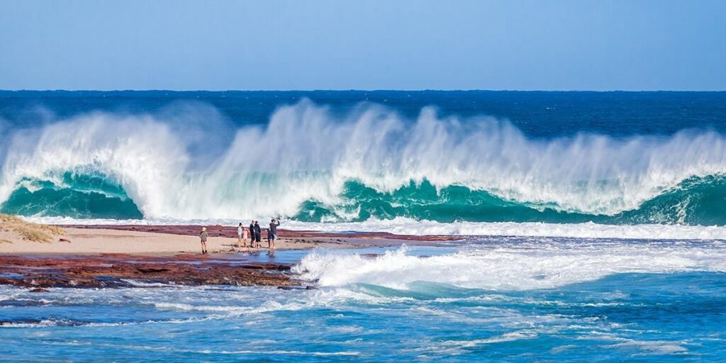 Visitors standing on the Kalbarri rock platform as large turquoise waves crash dramatically in the background.