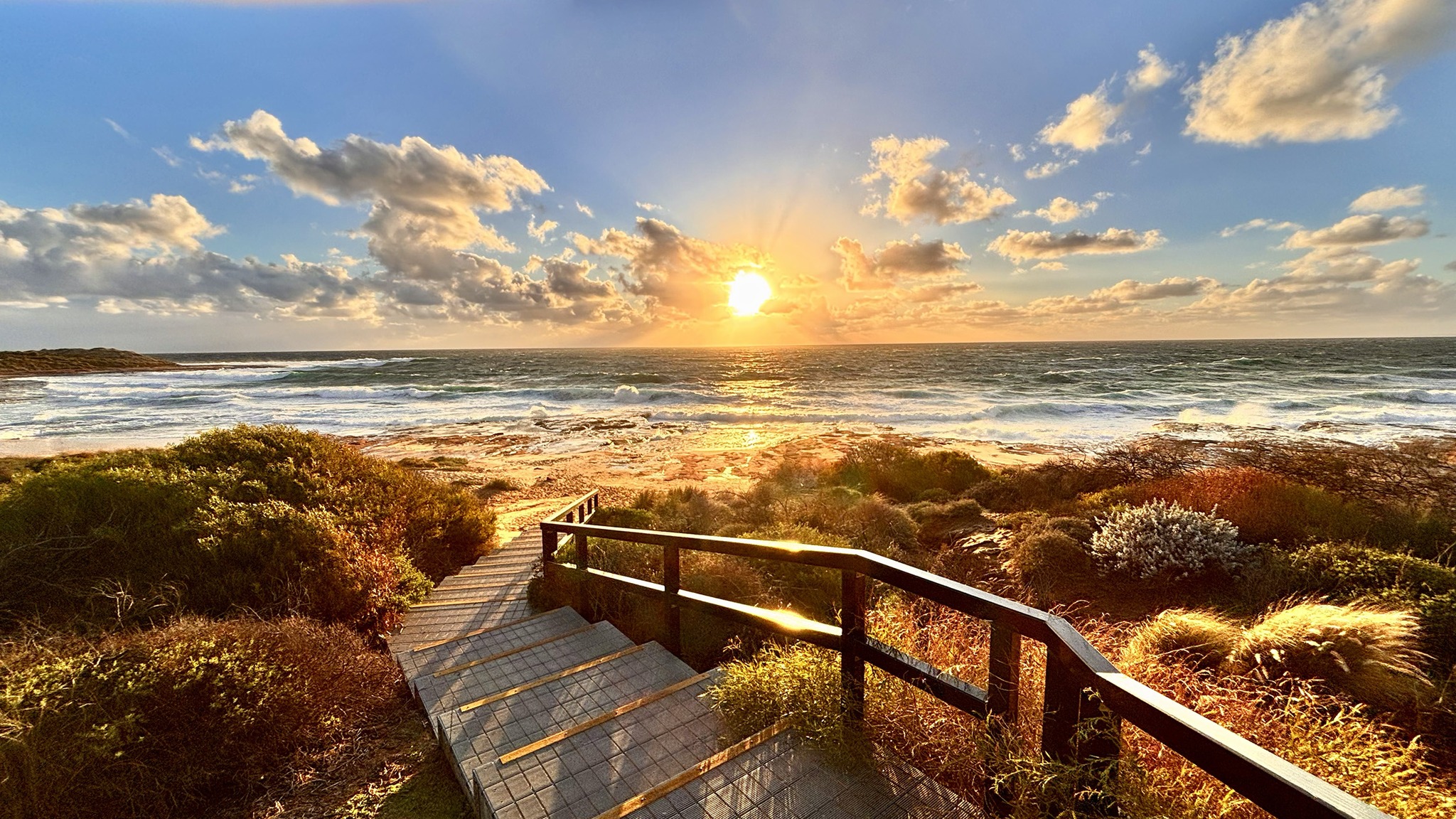 Sunset over the Indian Ocean from the Jakes Point boardwalk in Kalbarri, Western Australia, with golden light reflecting off the waves and native coastal vegetation.